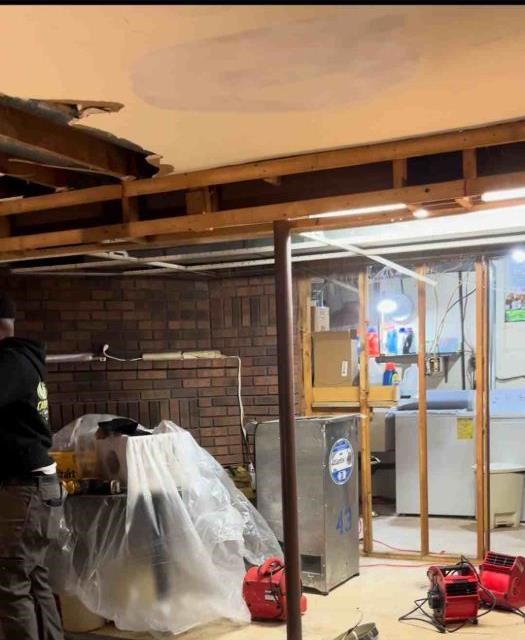 A basement mid-drying with the water-stained ceiling opened to the joists, contents sheeted in plastic, and air movers running on the floor