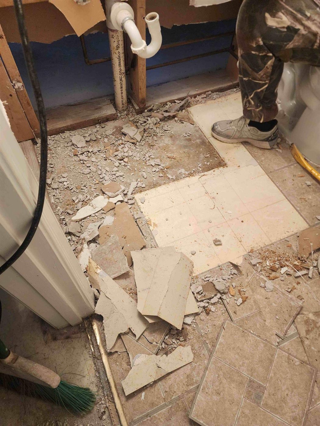 A bathroom floor being stripped back during a water damage job, with tile broken out to the subfloor and the wall opened to reach the plumbing
